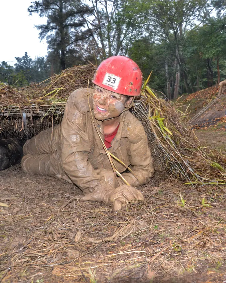 Andresa rastejando na lama sob uma rede, de capacete, sorrindo, durante treinamento.