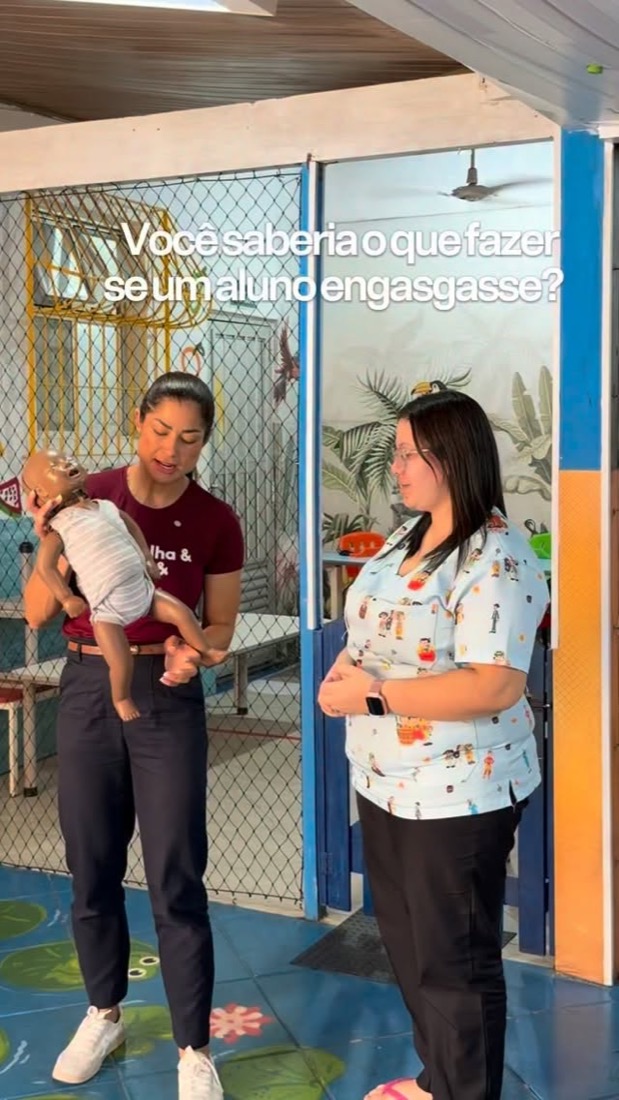 Andresa numa escola, ensinando primeiros socorros a uma professora com um manequim de bebê.