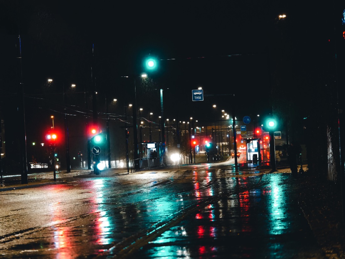 Avenida urbana à noite, com asfalto molhado e semáforos acesos.