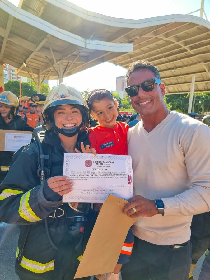 Andresa de equipamento completo de combate a incêndio, com o certificado do curso na mão, ao lado da família.