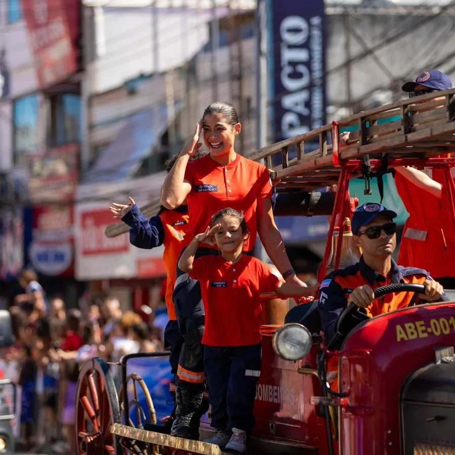 Andresa batendo continência em cima do carro antigo dos bombeiros, durante o desfile, com a rua cheia.