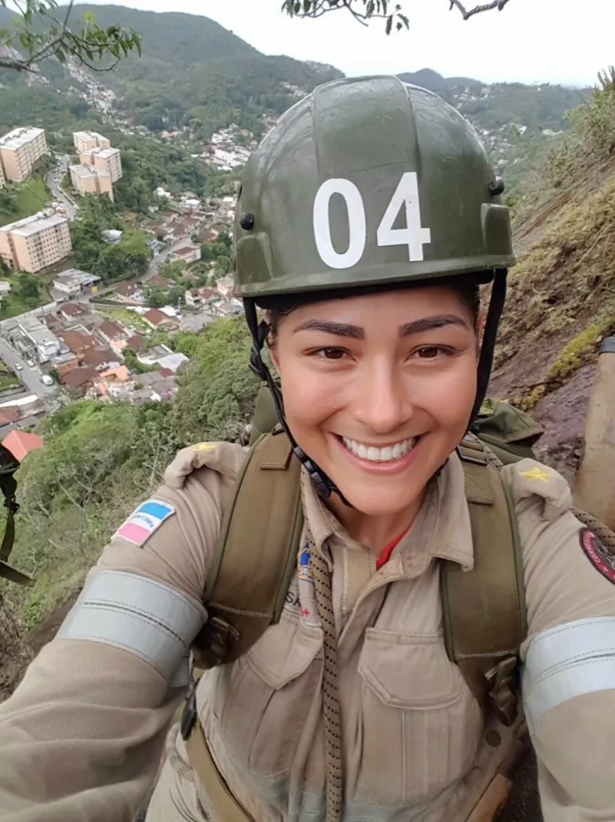 Andresa de capacete e equipamento de resgate, pendurada na encosta, com a cidade lá embaixo.